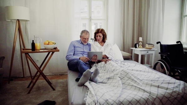 A senior couple with tablet at home, a husband and his wife sitting on bed, spending time together. Slow motion.