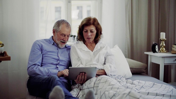 A senior couple with tablet at home, a husband and his wife sitting on bed, spending time together. Slow motion.