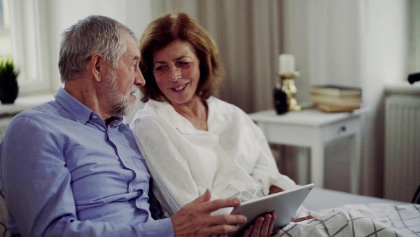 A senior couple with tablet at home, a husband and his wife sitting on bed, spending time together. Slow motion.