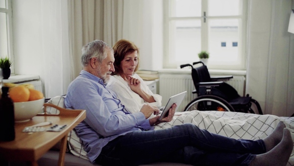 A senior couple with tablet at home, a husband and his wife sitting on bed, spending time together. Slow motion.