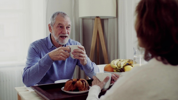 A senior couple with coffee sitting at the table at home, having breakfast and talking.