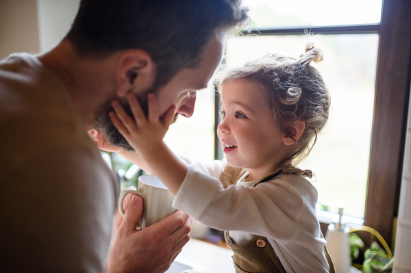 Father giving drink to sick toddler daughter indoors in kitchen at home.