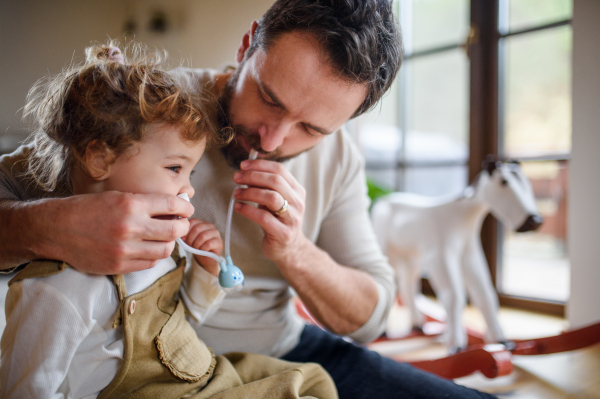 Mature father with small sick toddler daughter indoors at home, using nasal aspirator.