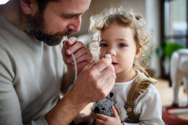 Mature father with small sick toddler daughter indoors at home, using nasal aspirator.