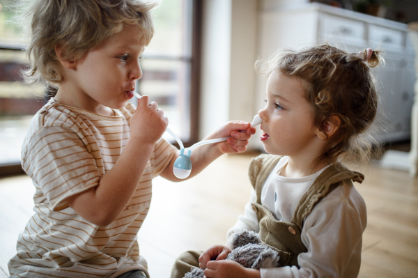 Two small children brother and sister indoors at home, playing with nasal aspirator.