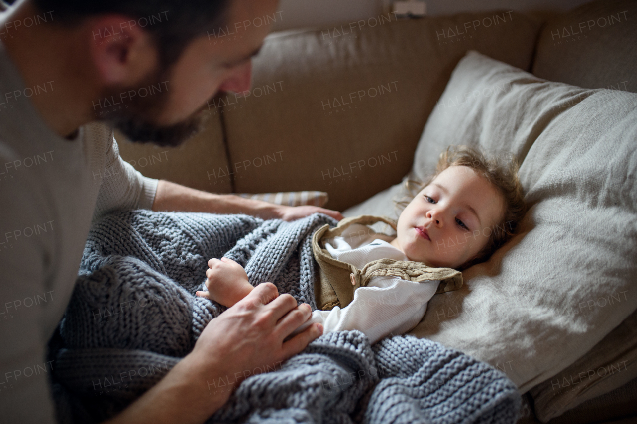 Unrecognizable father checking on small sick daughter lying in bed indoors at home.