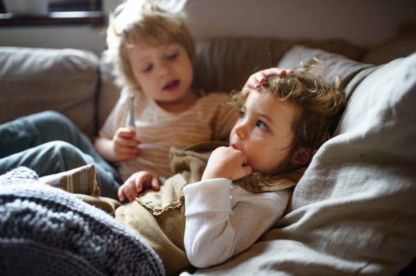 Two small sick children brother and sister at home lying in bed, measuring temperature.