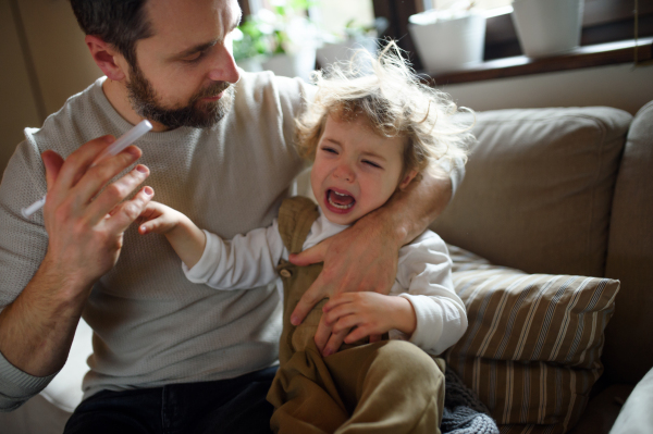 Front view of mature father giving syrup to small sick daughter indoors at home.