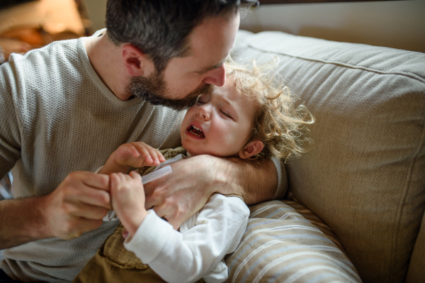 Father with small sick crying toddler daughter indoors at home, applying medication.