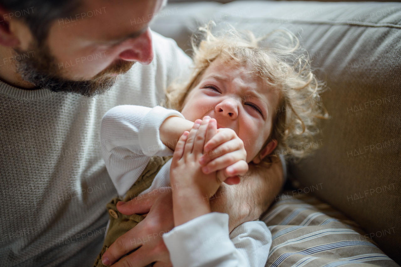 Mature father with small sick crying toddler daughter indoors at home.