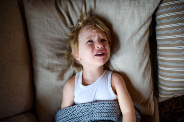 Top view of crying small sick boy lying in bed indoors at home.