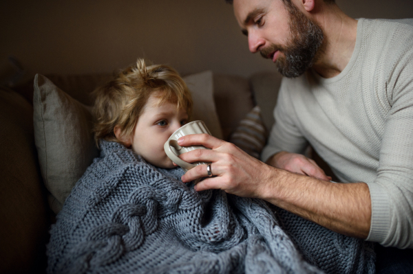 Front view of father giving drink to small sick son lying in bed indoors at home.