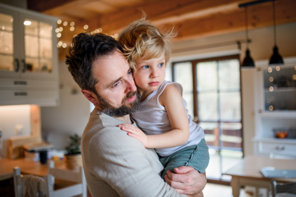 Father with small sick unhappy son indoors at home, holding and comforting him.