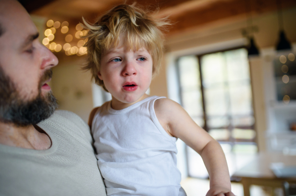 Father with small sick unhappy son indoors at home, holding and comforting him.