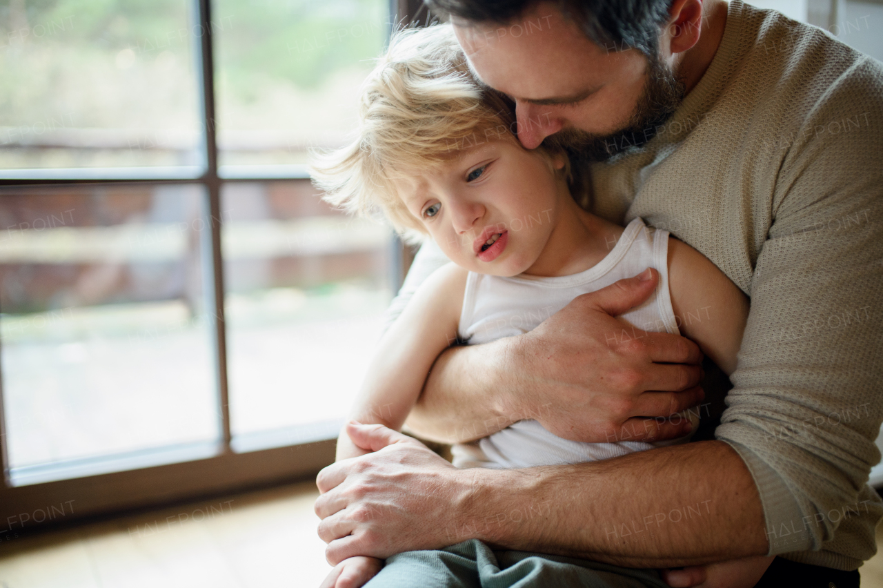 Father with small sick unhappy son indoors at home, holding and comforting him.