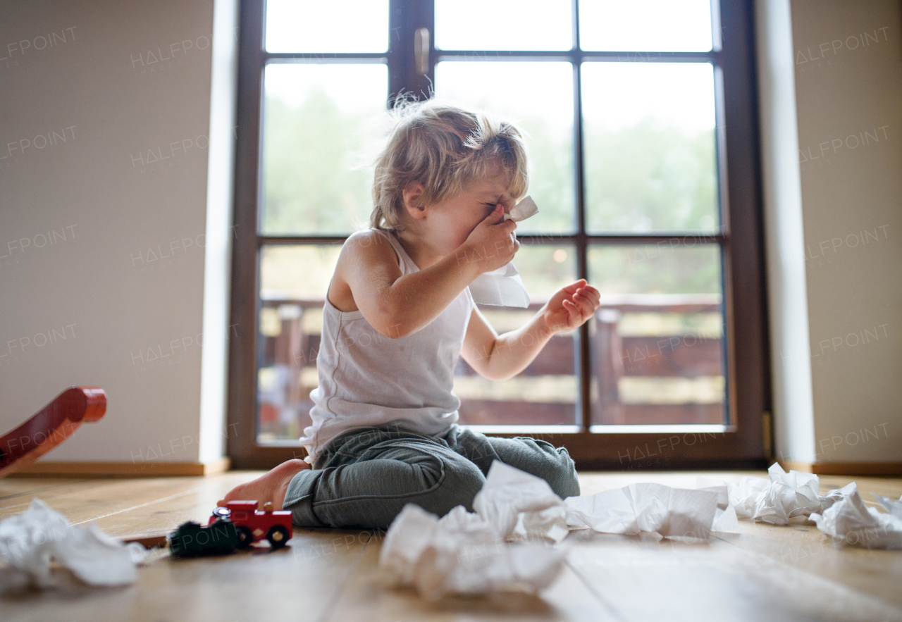 Small sick boy with cold at home sitting on floor, sneezing and blowing nose.