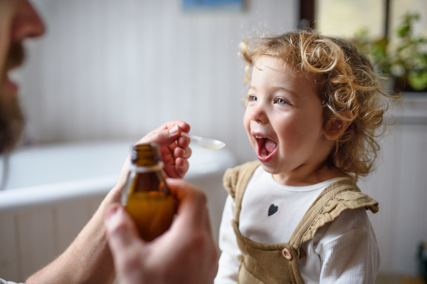 Unrecognizable mature father givingsyrup to small sick daughter indoors at home.