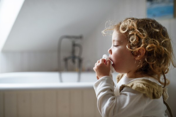 Side view of small sick girl with cold at home sitting on ground, sneezing. Copy space.