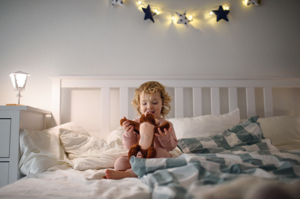 Small toddler girl sitting on bed indoors at home, playing with soft toy.