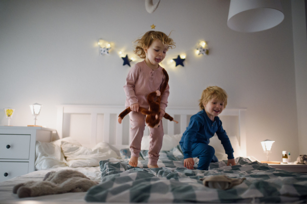 Two small laughing children jumping on bed indoors at home, having fun.