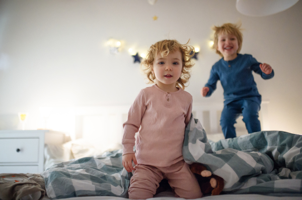 Two small laughing children jumping on bed indoors at home, having fun.