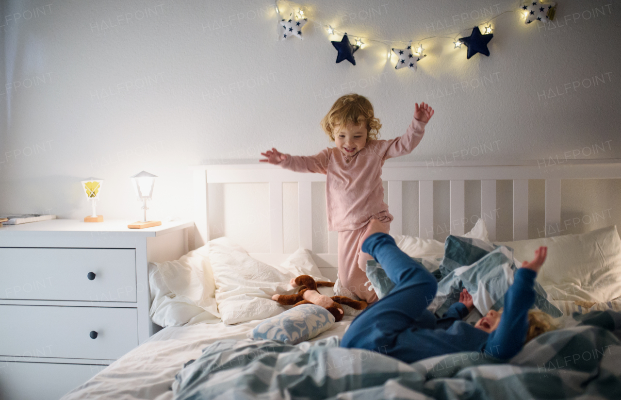 Two small laughing children jumping on bed indoors at home, having fun.