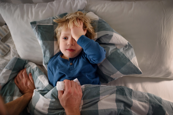 Unrecognizable father checking on small sick son lying in bed indoors at home.