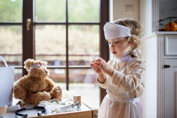 Small toddler girl with doctor uniform indoors at home, playing with teddy bear.