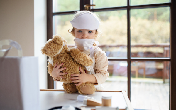 Small toddler girl with doctor uniform indoors at home, playing with teddy bear.