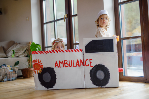 Two small children with doctor uniforms and toy ambulance car indoors at home, playing.