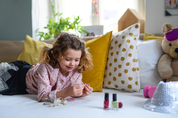 Happy small girl indoors at home, playing on bed with accessories. Lockdown concept.