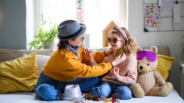 Portrait of sisters indoors at home, playing on bed. Lockdown and dressing up concept.