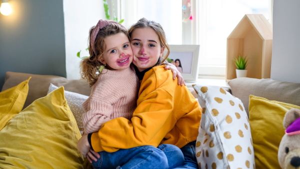 Portrait of sisters indoors at home, playing with lipstick. Lockdown concept.