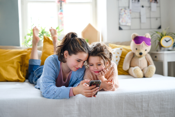 Portrait of sisters indoors at home, using smartphone. Lockdown concept.