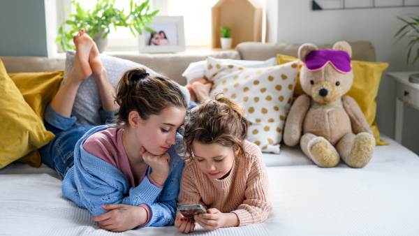 Portrait of sisters indoors at home, using smartphone. Lockdown concept.