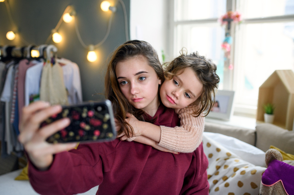 Portrait of sisters indoors at home, taking selfie with smartphone. Lockdown concept.