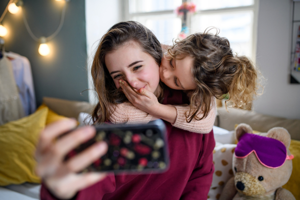 Portrait of sisters indoors at home, taking selfie with smartphone. Lockdown concept.