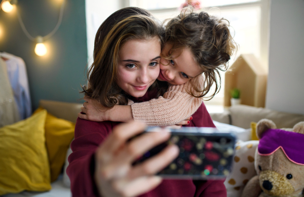 Portrait of sisters indoors at home, taking selfie with smartphone. Lockdown concept.