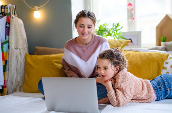 Portrait of sisters indoors at home, using laptop. Lockdown concept.