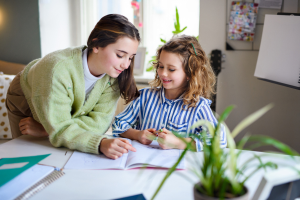 Portrait of teenager girl helping small sister to learn indoors at home, coronavirus and distance learning concept.