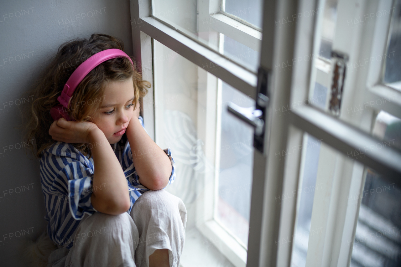 Portrait of sad small girl with headphones indoors at home, sitting on window sill.