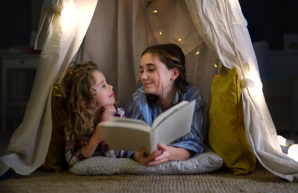 Sisters indoors at home, reading book in pajamas in tent. Lockdown concept.