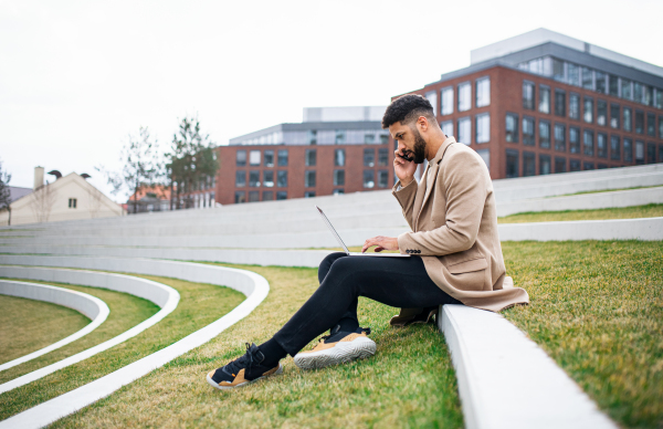 A man with laptop and smartphone outdoors in park in city, working.