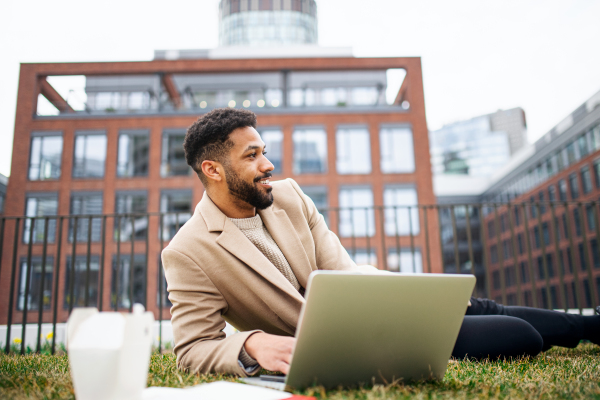 Young man with laptop working outdoors in park in city, remote office concept.