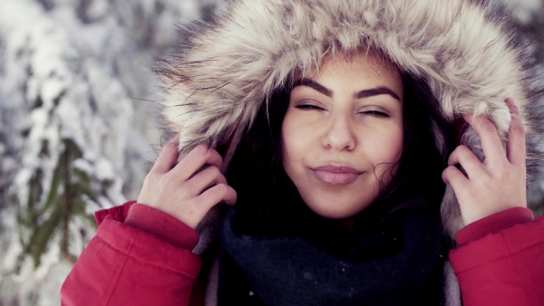 A front view portrait of young woman standing outdoors in snowy winter forest, wearing fur hood. Slow motion.