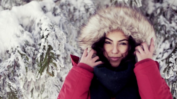 A front view portrait of young woman standing outdoors in snowy winter forest, wearing fur hood. Slow motion.