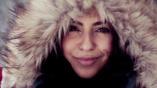 A front view portrait of young woman standing outdoors in snowy winter forest, wearing fur hood. Slow motion.