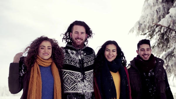 A group of young cheerful friends standing outdoors in snow in winter forest, looking at camera. Slow motion.