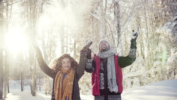 A portrait of couple standing outdoors in snow in winter forest, throwing snow. Slow motion.