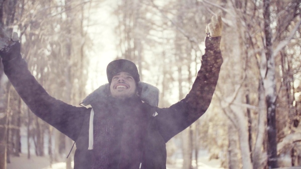 A portrait of young man standing outdoors in snow in winter forest, throwing snow. Slow motion.